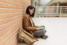 Positive Female Student Sitting On Floor And Reading Book While Studying In University Corridor