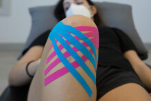 Crop Anonymous Female Athlete In Mask With Elastic Kinesio Tapes On Leg Lying On Medical Bed While Looking Away In Physiotherapy Center