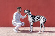 © Sergio Victor Vega/ADDICTIVE STOCK - Side view of young bearded guy in stylish outfit sitting on haunches and stroking obedient Harlequin Great Dane dog against red wall on street