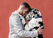 © Sergio Victor Vega/ADDICTIVE STOCK - Side view of happy young unshaven male in casual clothes and adorable obedient Harlequin Great Dane dog hugging each other against red background