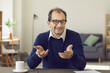 © Studio Romantic - Self presentation at offline or online conference meeting concept. Portrait of smart smiling mature man in eyeglasses with gesturing hand sitting at table looking at camera. Home office interior