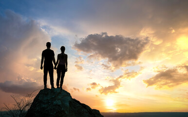 Wall Mural - Man and woman hikers standing on a big stone at sunset in mountains. Couple together on a high rock in evening nature. Tourism, traveling and healthy lifestyle concept.