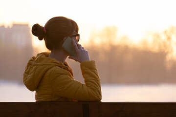 Young pretty woman sitting on a park bench talking on her smartphone outdoors in warm autumn evening.