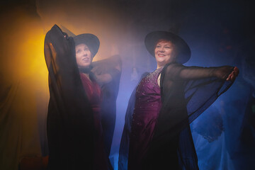 Two adult women looking as witches having fun and posing in dark room decorated for Halloween during photoshoot in studio.