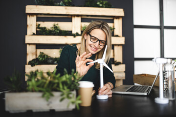 pretty, young and blond woman with stylish, modern black glasses sits in a sustainable office and works with pinwheel wind turbine models