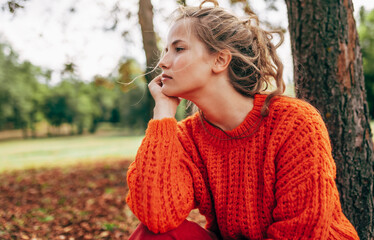 Caucasian young woman resting in the park. Female wearing an orange knitted sweater has pensive expression posing on the nature background.