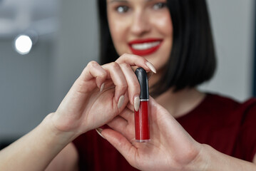 Canvas Print - Candid portrait of a pretty young woman with red lips smiling broadly showing her favorite lipstick in focus. Gorgeous brunette female with red lips making an online tutorial with beauty product.