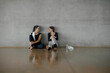 © BASILICOSTUDIO STOCK - Smiling sporty young women resting after workout. Female athletes sitting against a wall of a big gym with wide gray concrete wall, in sportswear, taking a break, talking and having fun. Copy space.