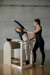 © BASILICOSTUDIO STOCK - Fit brunette female coach helping a woman to practice Pilates stretching exercise in a ladder barrel equipment, combo chair, during personal fitness lessons in a fitness center gym, health club.