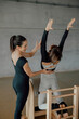 © BASILICOSTUDIO STOCK - Portrait of Beautiful brunette female coach helping a woman to practice Pilates stretching exercise in a ladder barrel equipment, combo chair, during personal fitness lessons in a fitness center gym.