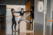 © BASILICOSTUDIO STOCK - Fit Woman doing Pilates exercise and postures on the ladder barrel equipment, helped for a female personal trainer, they wear protective masks, in a big loft fitness club with natural light.