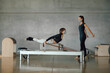 © BASILICOSTUDIO STOCK - Caucasian Brunette Fitness coach with face mask, giving instructions to a sporty woman during a Pilates lesson, while pulling a stretch bands from a Reformer Machine. Prevention during Covid-19.