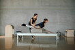 © BASILICOSTUDIO STOCK - Full shot of Woman doing core exercises during Personal Pilates class on a Reformer machine, by a female instructor at a big gymnasium center, wide industrial studio. Healthy fitness lifestyle.