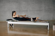 © BASILICOSTUDIO STOCK - Young caucasian woman doing exercises on fitness machine in gym. Female using rowing Pilates machine at fitness club, practicing with resistance bands on reformer. Indoors daytime.
