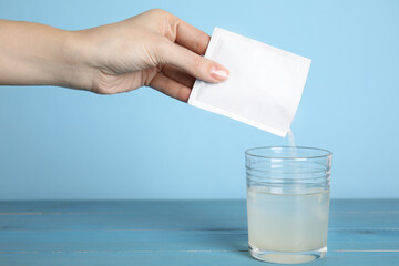 Poster - Woman pouring powder from medicine sachet into glass of water on blue wooden table, closeup