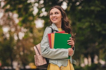 Poster - Photo of pretty cute young woman dressed teal jacket glasses backpack holding colorful copybooks looking back outdoors city park