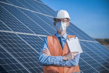 Wall Mural - Engineer, man in uniform and mask, helmet glasses and work jacket on a background of solar panels at solar station. Technician check the maintenance.