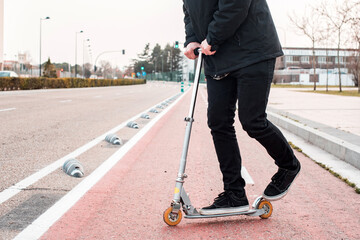 Wall Mural - Detalle de un hombre joven vestido con ropa negra de invierno montando en patinete por un carril bici de la ciudad. Scooter no eléctrico. Concepto de transporte sostenible