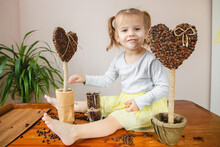 A Little Girl With Two Ponytails Sits On A Table Next To Self-made Coffee Heart-shaped Trees