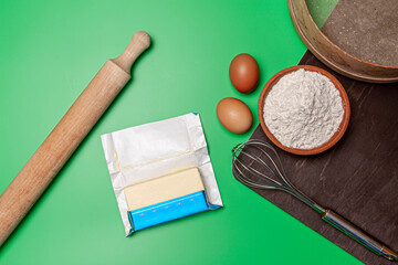 a wooden rolling pin, two eggs, wooden bowl filled with flour, sieve, stick of butter and black cutting board on green background