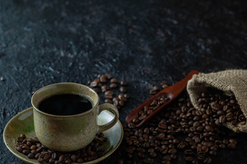 Hot black coffee for morning beverage menu in vintage brown ceramic cup with coffee beans roasted in burlap sack bag on dark grunge rustic table background. Flat lay with copy space.