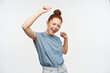 Portrait of attractive, happy redhead girl with hair gathered in a bun. Wearing blue t-shirt and jeans. Excited. Lifts her arms and dance with closed eyes. Stand isolated over white background