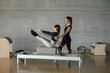© BASILICOSTUDIO STOCK - Slim fit Woman doing core exercises while taking Personal Pilates lesson on a Reformer machine, by a female instructor at a big gymnasium center, wide industrial studio. Healthy fitness lifestyle.