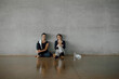 © BASILICOSTUDIO STOCK - Smiling sporty young women take a break after workout. Confident Female instructors sitting and leaning against wall in sportswear, looking at camera, in a wide gymnasium, gray wall, copy space.