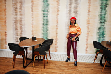 Wall Mural - Elegant afro american woman in red french beret, big gold neck chain polka dot blouse and leather pants pose indoor.