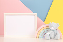 White Blank Wood Frame With Teddy Bear And Pastel Toy Rainbow On White Desk. Blank Horizontal Frame Mockup, Baby Room Art With Baby Kid Toys Over Pink, Yellow And Blue Background