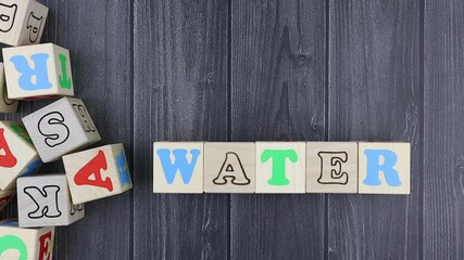 Fototapete - Colourful wooden toy alphabet blocks spelling word - Water over a dark grey plank background with copyspace. Stop motion, top view