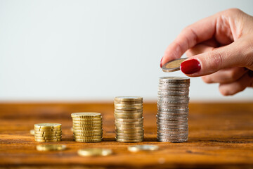 Female hand putting two euros onto stack on wooden table. Concept of pile of coins increasing by saving salary. Womans finger holding metal money.