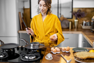 Young woman dressed in yellow bathrobe making breakfast, frying eggs on a cooking pan on the kitchen at home