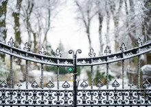 Old Iron Gates With Victorian Ironwork Locked Closed With Snowy Scene Falling And Heavy Snow Settling On Metal.  Line Of Trees In Winter Landscape Entrance Driveway.