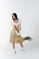 Sticker - full length of happy woman in spring outfit smiling while holding straw bag with flowers on white