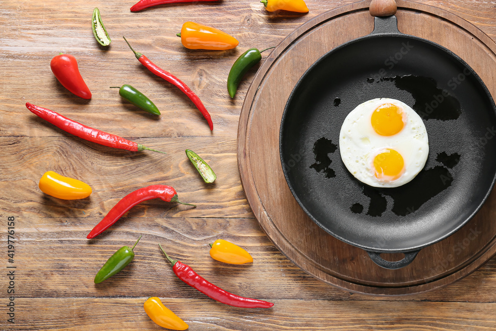 Frying pan with tasty eggs and chili peppers on wooden background