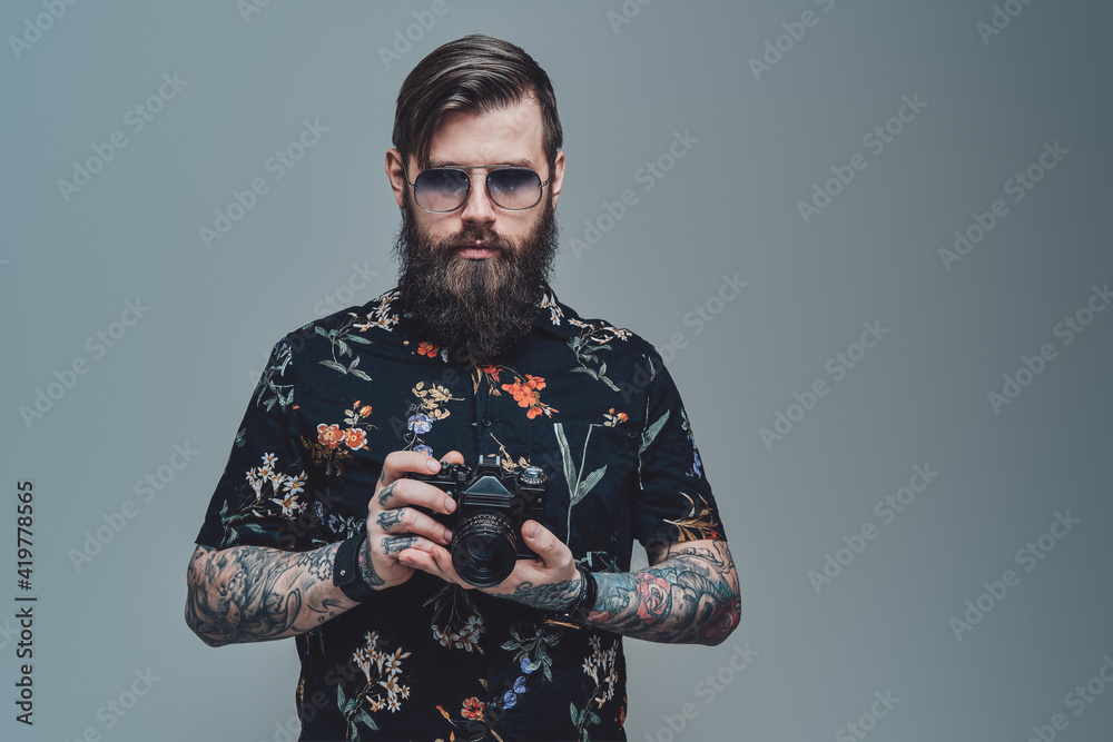 Successful hipster person with bearded face and modern coiffure posing in gray background holding photo camera. - obrazy, fototapety, plakaty 