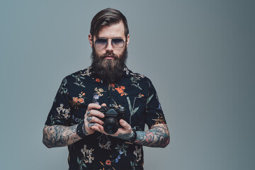 Successful hipster person with bearded face and modern coiffure posing in gray background holding photo camera.