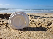 Single-use White Plastic Cup Lid Cover On The Summer Sandy Beach In Front Of The Water Ocean. Dirty Beach With Trash.