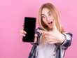 © Studio Nova - Surprised happy blonde woman in shirt showing blank smartphone screen and pointing at it while looking at camera with open mouth on pink background