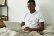 © Anatoliy Karlyuk - Concentrated young black businessman has to stay home because of seasonal flu, working distantly from bed using wireless high speed internet connection on portable computer, having serious look