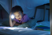 Asian Preschool Boy Child To Make A Camp To Play Imaginatively And Reading Book By Flashlight  In Living Room At Home..