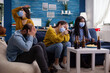© DC Studio - Disappointed diverse group of people playing video games respecting social distancing because of orona outbreak wearing face mask not to spread the virus and get sick. Beer bottle and chips.