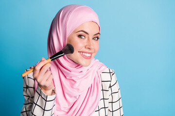 Wall Mural - Close-up portrait of pretty cheerful muslimah girl applying decorative rouge isolated over bright blue color background