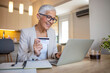 © Dragana Gordic - Shot of a senior woman using a credit and mobile while working on her finances at home. Senior woman shopping online. Happy senior woman making online payments of bill using laptop.