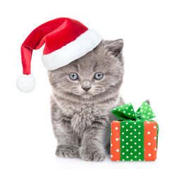 Canvas Print - Cute kitten wearing a red santa hat sits with gift box and looks at camera. isolated on white background