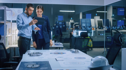 Factory Office Meeting Room: Male Chief Engineer Holds Digital Tablet Computer, Shows Screen to Female Project Manager, They Talk, Find Optimization Solution. Modern Industry 4.0 CNC Machinery