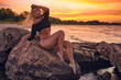© aleonovs - Young beautiful adult sexy blonde model pose in the black bikini by sitting on the rocks and taking fun at dramatic sunset