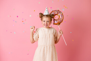 A happy little girl in a birthday cap holds a doughnut-shaped balloon on a pink background in the studio, space for text