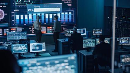Poster - Two Software Engineers Talking Next to a Big Screen in a Modern Monitoring Office with Live Analysis Feed with Charts. Monitoring Room Big Data Scientists and Managers Sit in Front of Computers.